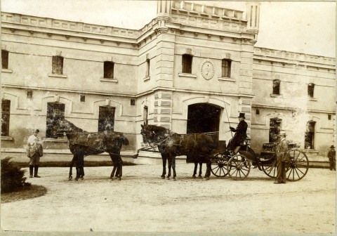 Wejście główne do pałacu w Będlewie widzianego w dzień. Przed wejściem dwa zaprzęgi konne na ścieżce z woźnicą, koniuszym i panem w kapeluszu. Woźnica trzyma bat w ręce.