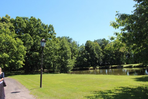 Fragment ścieżki, na której stoi kobieta, trawy z latarnią i stawu zacienionego drzewami w parku przy pałacu w Kobylnikach widziane w dzień.